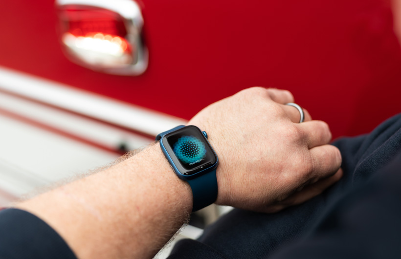 Cerca de un camión de bomberos, un brazo con un Apple Watch que muestra el mensaje "Ahora inspira…"